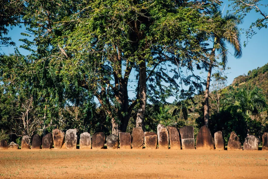 A row of upright carved stone monoliths stands on a grassy clearing at Parque Ceremonial Caguana, surrounded by lush tropical trees and hills under a clear blue sky.