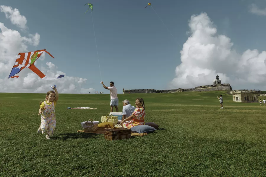 A family enjoys a picnic on a grassy hill, flying colorful kites from the local brand Chiringas.com under a blue sky with fluffy clouds. The historic structure of El Morro is visible in the background.