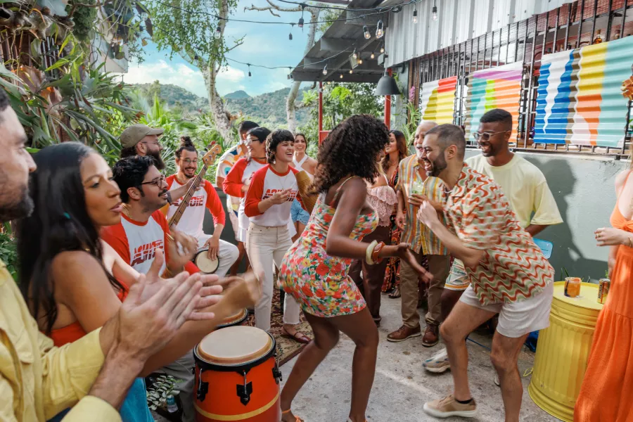 A lively outdoor gathering with people dancing to live music, playing drums and guitar, and celebrating together on a colorful patio surrounded by tropical plants and mountain views.