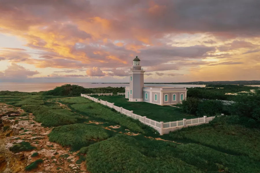 Los Morrillos Lighthouse in Cabo Rojo stands on a grassy coastal cliff, surrounded by a white fence, overlooking the ocean under a colorful sunset sky with orange and pink clouds.
