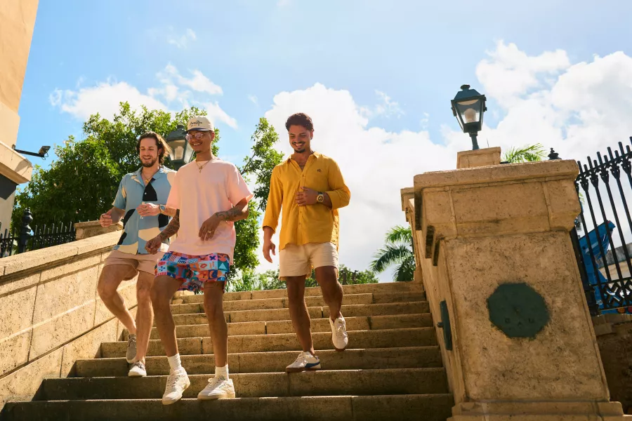 Three men wearing relaxed, colorful clothing walking down stone steps under a bright sky in Old San Juan