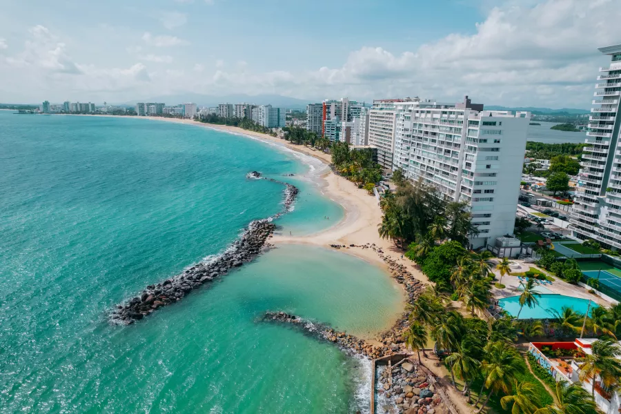 Aerial view of Isla Verde shoreline in Puerto Rico, featuring turquoise ocean waters, sandy beaches, and a line of high-rise beachfront hotels and palm trees along the coast, with rocky breakwaters extending into the sea under a partly cloudy sky.