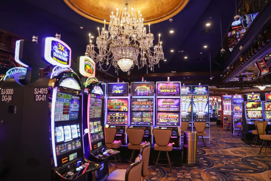 A row of glowing slot machines at Foxwoods El San Juan Casino, featuring vibrant digital screens under a magnificent, large crystal chandelier and a deep blue ceiling.