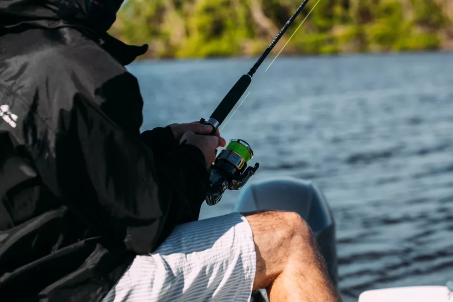 Close-up of a person fishing from a boat in calm coastal waters, holding a fishing rod and reel near the shoreline