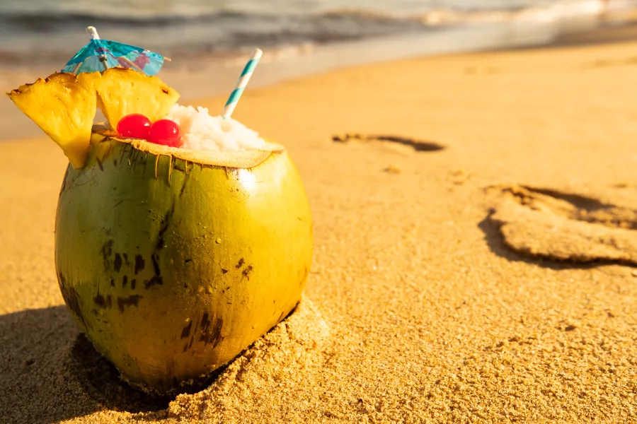 A tropical Piña Colada served in a fresh green coconut on a sun-drenched sandy beach, garnished with pineapple slices, cherries, and a blue umbrella with the ocean waves in the blurred background.