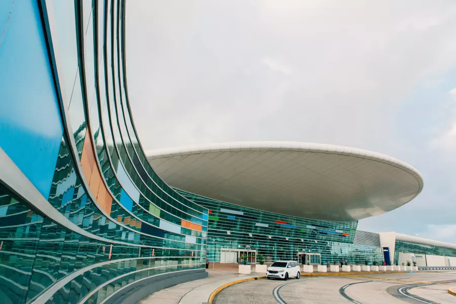 The modern exterior of Terminal A at the Luis Muñoz Marín International Airport (SJU) in San Juan, featuring a dramatic curved glass wall with colorful panels and a large, white circular canopy roof.