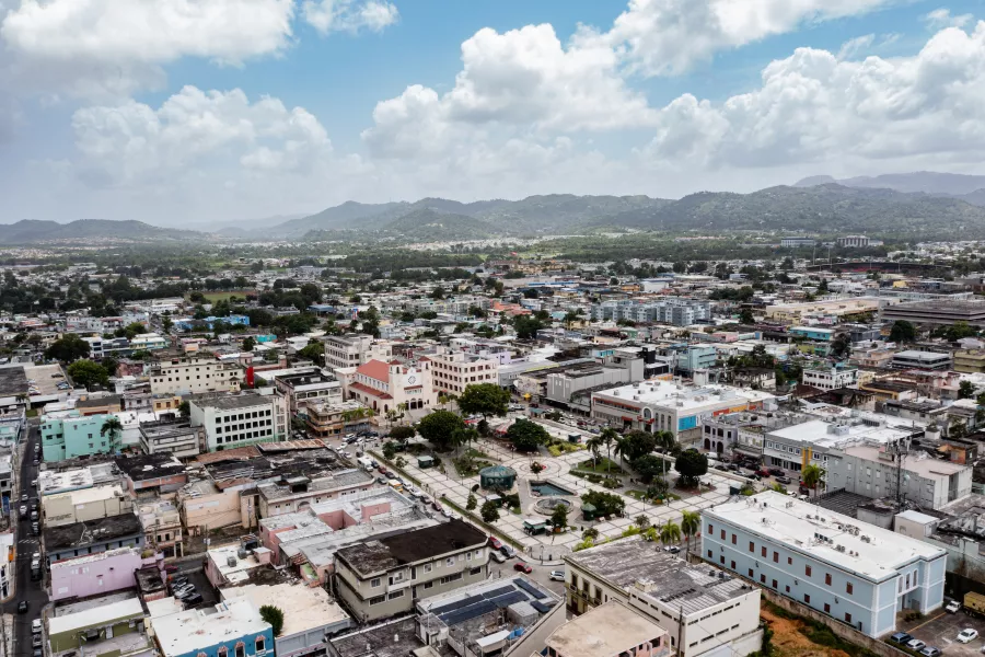 Aerial view of downtown Caguas, Puerto Rico, with colorful buildings, a central plaza, and green mountains in the background under a bright blue sky.