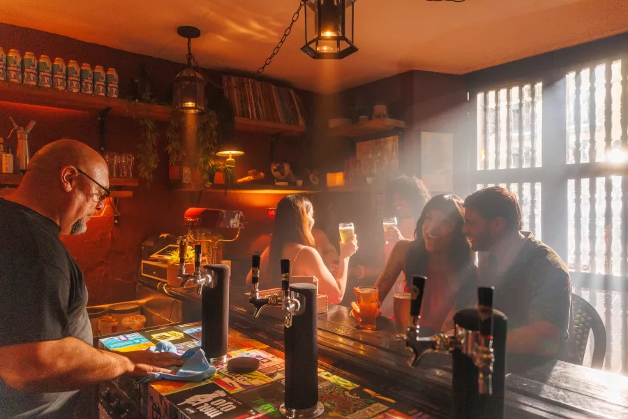 Travelers enjoy drinks at a cozy Old San Juan bar as a bartender cleans the counter and sunlight streams through the windows.
