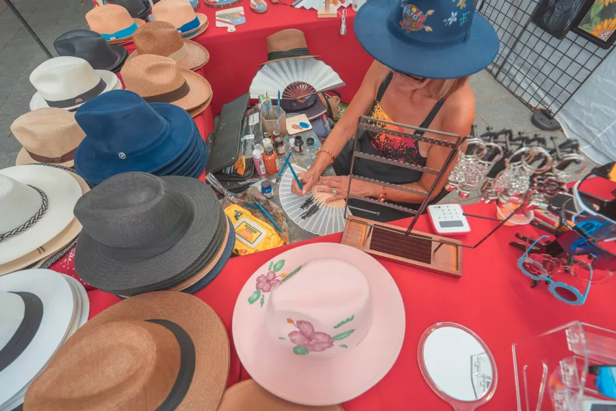 Una artesana pinta a mano un abanico decorativo en una feria al aire libre en Puerto Rico, rodeada de sombreros, joyería y otras artesanías locales.
