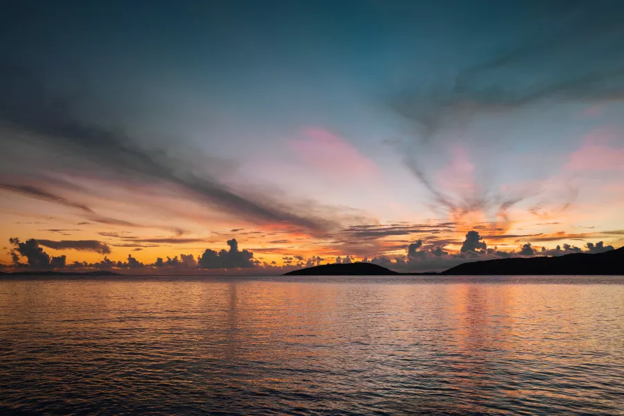 Sunrise over Playa Melones in Culebra, with calm waters reflecting shades of orange, pink, and blue beneath streaked clouds and the dark silhouettes of nearby cays along the horizon.