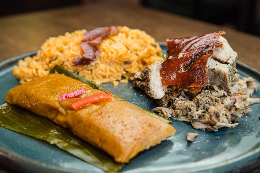Plate of traditional Puerto Rican cuisine featuring roasted pork with crispy skin, arroz con gandules, and a pastel wrapped in a plantain leaf.