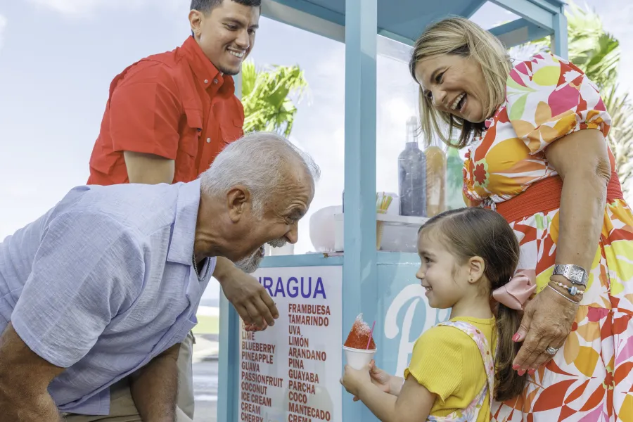A girl holds a red piragua beside two adults and a vendor at a blue shaved-ice cart.