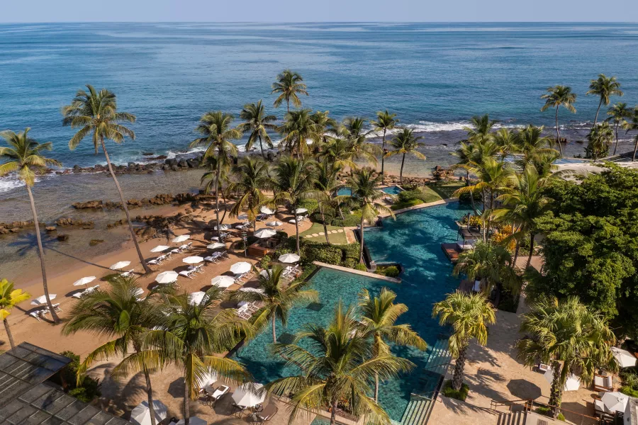 Vista aérea de Dorado Beach, a Ritz-Carlton Reserve, que muestra múltiples piscinas color turquesa rodeadas de exuberantes palmeras y jardines tropicales. Sillas reclinables y sombrillas blancas bordean una playa de arena junto a una tranquila costa rocosa, mientras el azul del océano Atlántico se extiende hasta el horizonte bajo un cielo despejado.