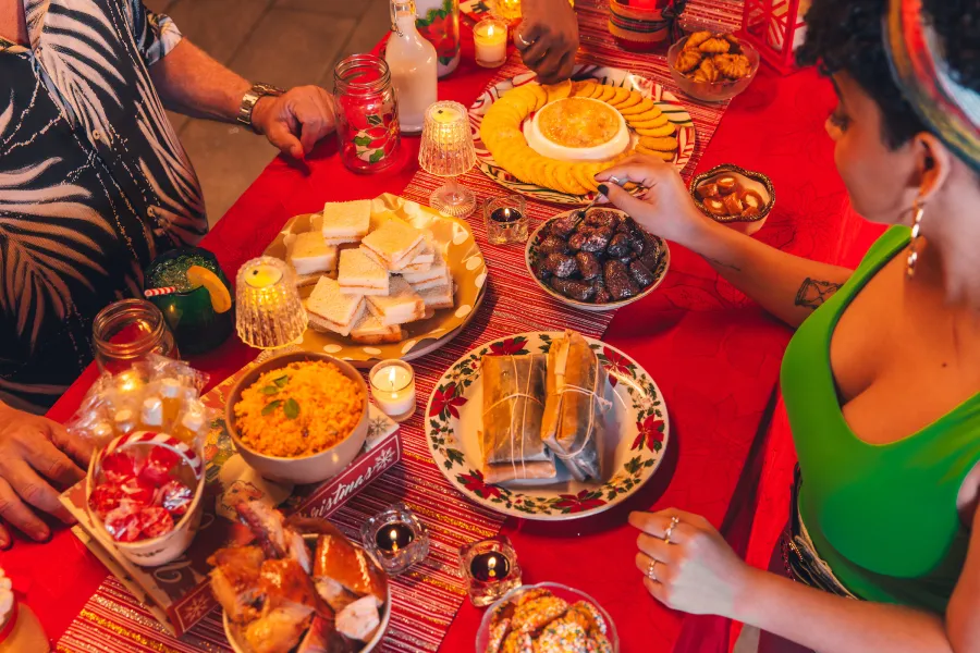 Las personas comparten un banquete navideño, que incluye pasteles envueltos, arroz, sándwiches y aperitivos, sobre una mesa roja decorada con velas.