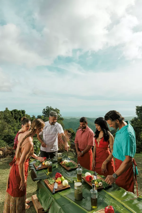 Group gathered around a chef preparing fresh local ingredients on a table covered with banana leaves, overlooking lush mountains and coastline in Puerto Rico.
