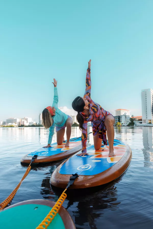 Two women practicing yoga on stand-up paddleboards in calm water with a city skyline in the background in Puerto Rico.