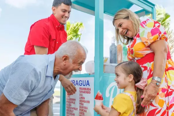Three generations of a family gather around a colorful piragua cart near Castillo San Felipe del Morro in Old San Juan, Puerto Rico, as a young girl enjoys a bright red shaved ice treat on a sunny day.