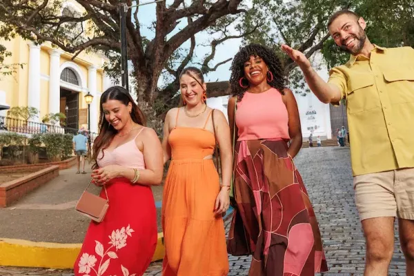 Four travelers walk along a cobblestone street in Old San Juan, Puerto Rico, smiling and enjoying a guided stroll beneath a large shade tree, with historic colonial architecture and street lamps in the background.