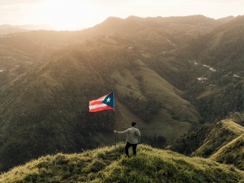 A man stands with a Puerto Rican flag atop a mountain summit as the sun sets.