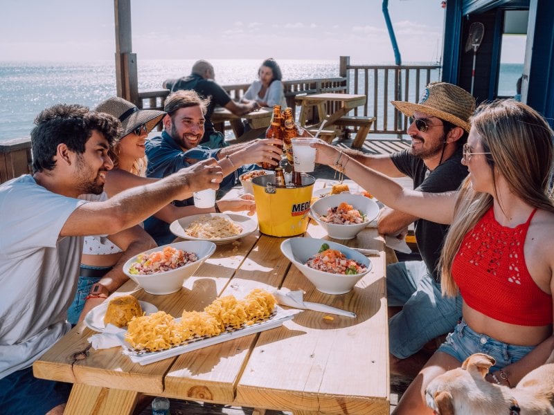 A group of friends toasts at an outdoor table overlooking the sea.
