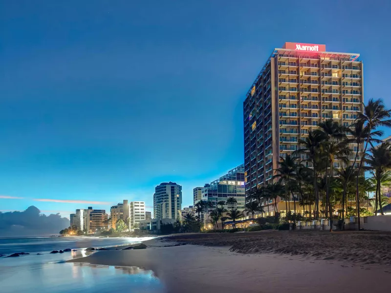 El exterior del San Juan Marriott Resort & Stellaris Casino, con la palabra "Marriott" iluminada, con la playa en primer plano y el cielo nocturno detrás.