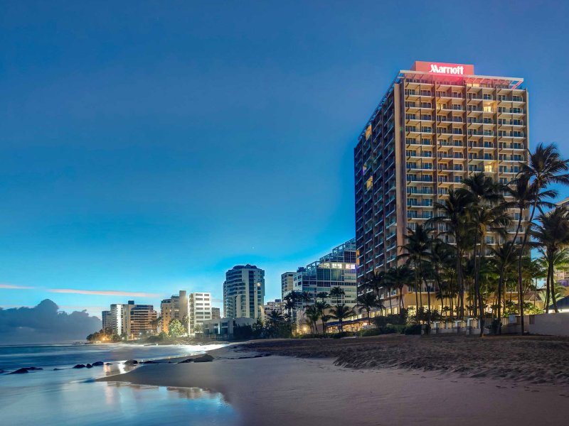 The exterior of the San Juan Marriott Resort & Stellaris Casino, with the word "Marriott" illuminated, with the beach in the foreground and the night sky behind it.