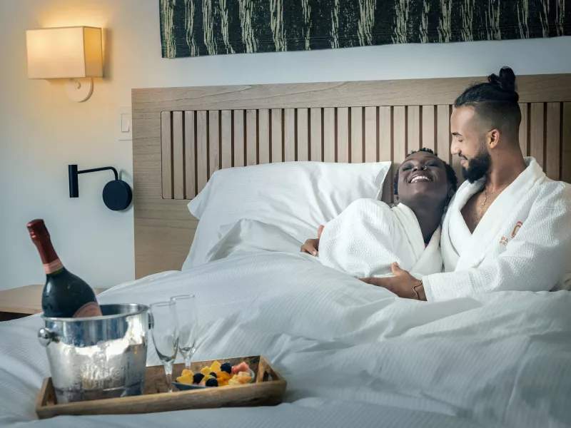 A couple lies in a hotel bed wearing white robes with a bottle of wine at the Hotel Palacio Provincial in San Juan, Puerto Rico.