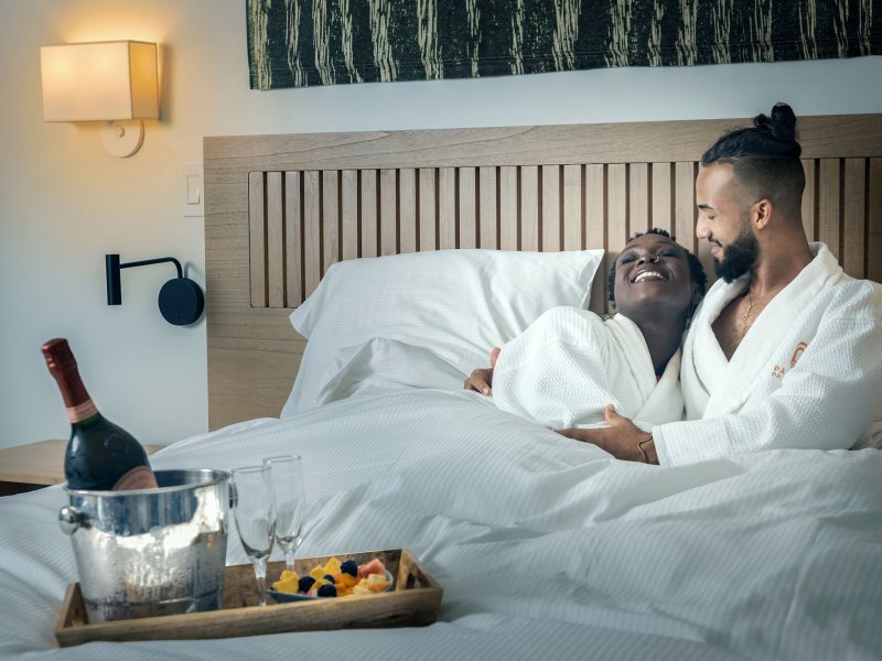 A couple lies in a hotel bed wearing white robes with a bottle of wine at the Hotel Palacio Provincial in San Juan, Puerto Rico.