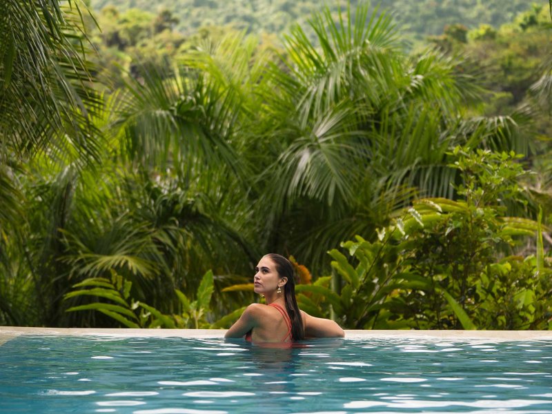 A woman rests on the side of an infinity pool overlooking a lush forest in the mountains of Puerto Rico.