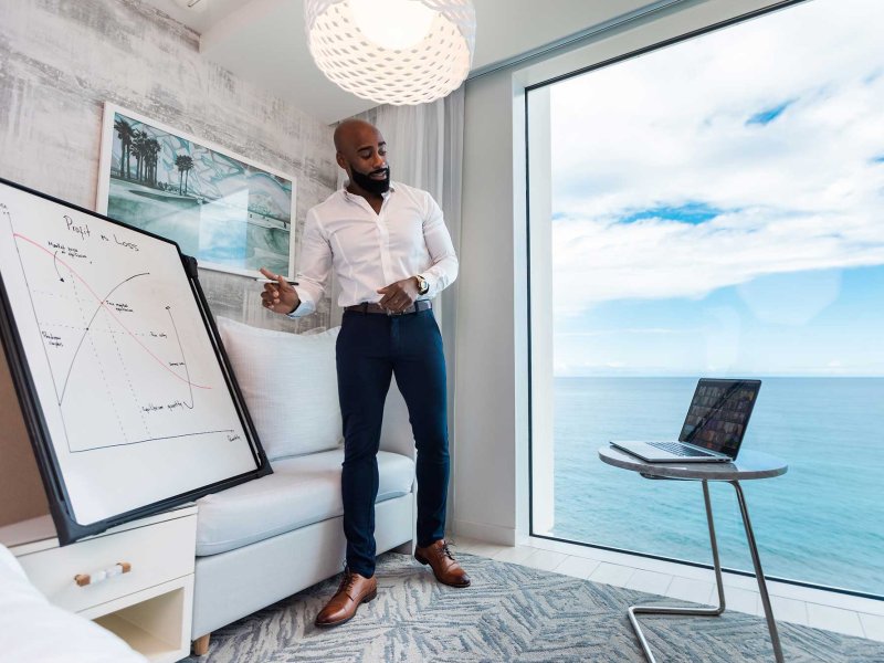 A man stands in front of a white board and a large window with an ocean view while making a remote presentation to his computer.