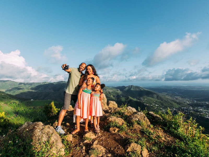 A mother, father, and two young girls pose for a selfie on top of a mountain with a dramatic backdrop in Puerto Rico.