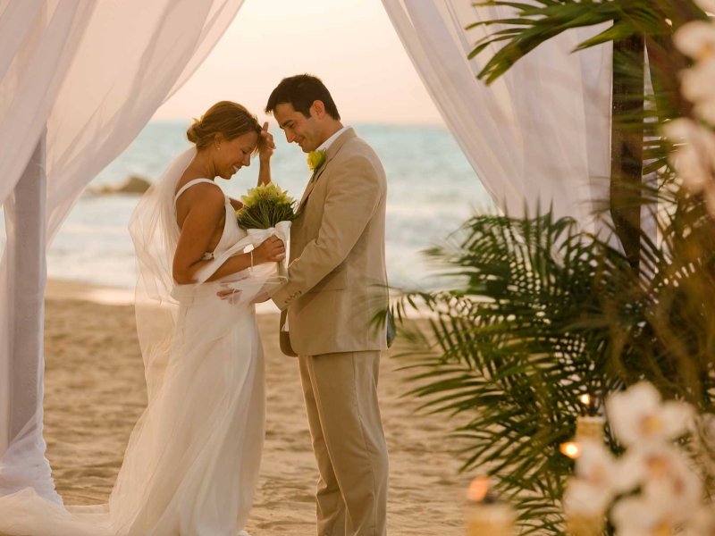 A bride and groom face each other in front of an altar draped with white fabric on a beach in Puerto Rico.