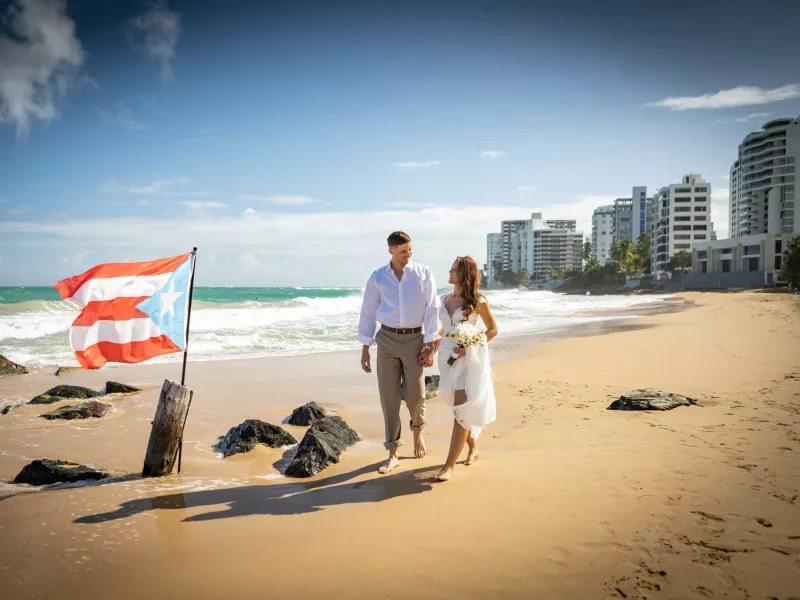 A bride and groom walk on the beach near the San Juan Marriott Resort & Stellaris Casino, with a Puerto Rican flag planted in the sand.