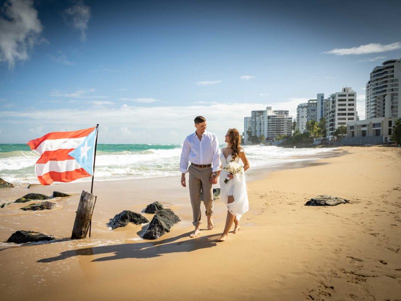 A bride and groom walk on the beach near the San Juan Marriott Resort & Stellaris Casino, with a Puerto Rican flag planted in the sand.