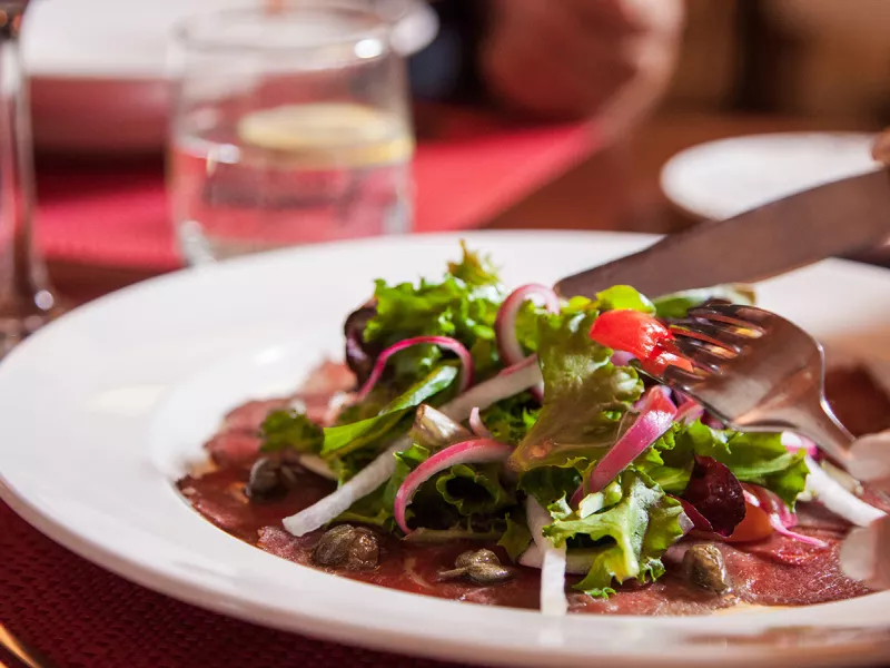 A person cutting a plate of salad greens on top of thinly sliced beef at Restaurante Alexandra.