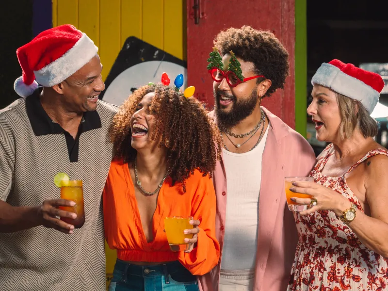 Four people laugh together in Santa hats and festive accessories, with three holding tropical drinks.