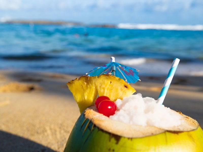 Foto de coco relleno de piña colada con una playa de fondo.