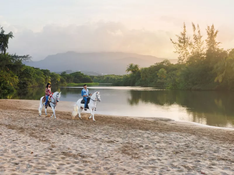 Two people riding white Paso Fino horses along a sandy riverbank at sunset, surrounded by tropical trees and mountains in Puerto Rico.