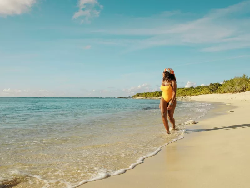Woman in a yellow swimsuit walking along a quiet sandy beach in Puerto Rico, with gentle waves washing ashore under a clear blue sky.