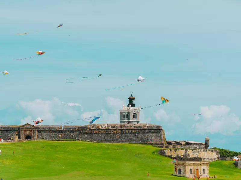 Colorful kites fly in a bright blue sky above El Morro fortress in San Juan, Puerto Rico, with its historic stone walls, lighthouse tower, and a wide green lawn in the foreground where a few people are scattered.