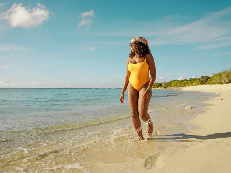 Woman walking along a beach wearing a yellow swimsuit and a colorful headscarf