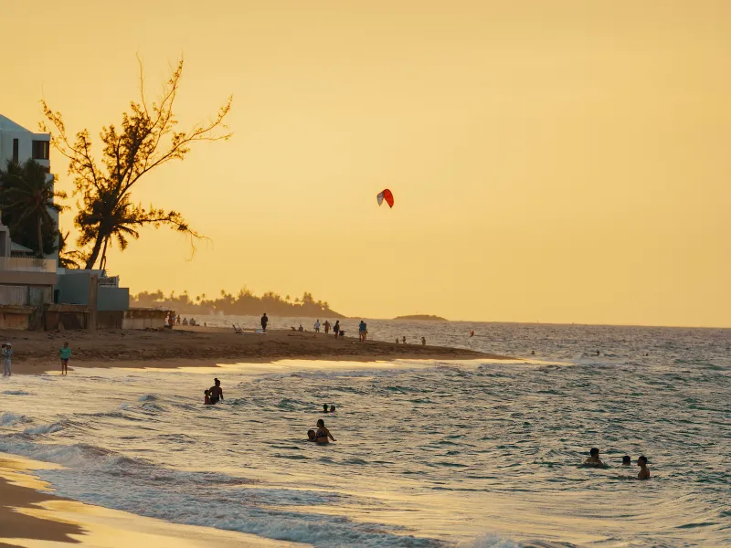 People enjoying the ocean at sunset on a beach in Puerto Rico, with a kite flying above the shoreline.