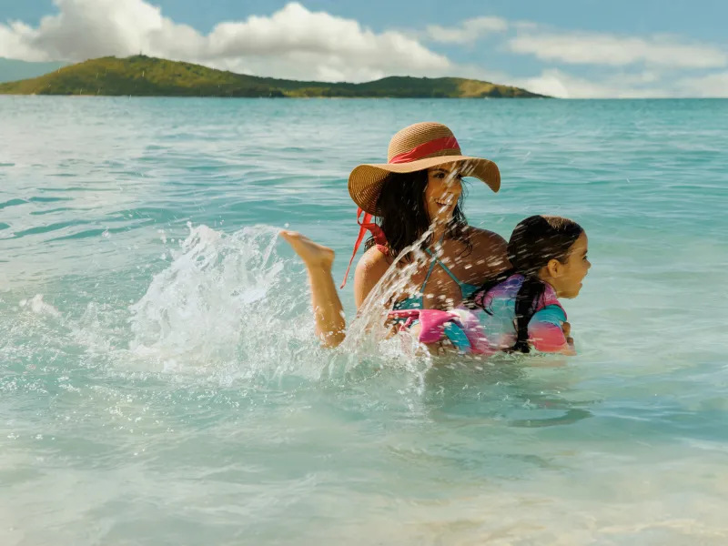 A female traveler with her kid enjoying the shallow turquoise waters of Cayo Icacos near Fajardo, with calm Caribbean seas, a sandy shoreline, and green hills in the distance.