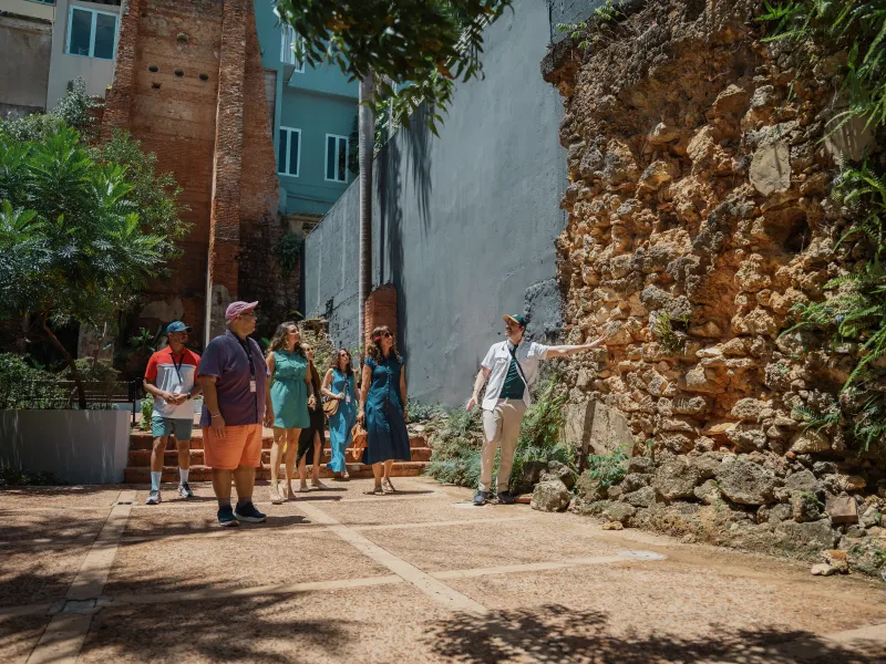 A tour guide points out historic stone ruins to a group visiting a courtyard in Old San Juan, Puerto Rico, surrounded by tropical greenery and colorful buildings.