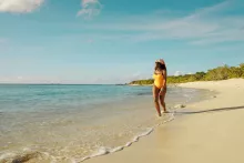Woman in a yellow swimsuit walking along a quiet sandy beach in Puerto Rico, with gentle waves washing ashore under a clear blue sky.