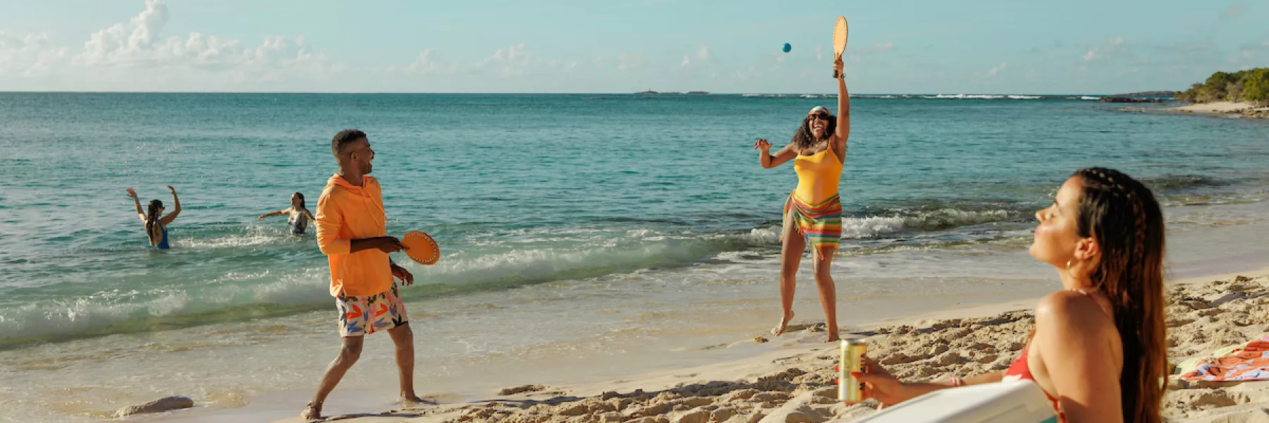 Friends playing paddleball and relaxing on a sandy beach along Puerto Rico’s east coast, with others swimming in the clear turquoise water under a sunny sky.