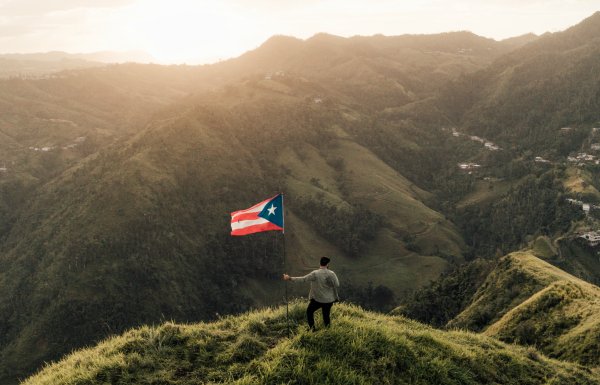 A man stands with a Puerto Rican flag atop a mountain summit as the sun sets.