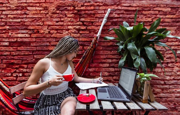 A woman works on her laptop computer at an outdoor desk against a vivid red brick wall.
