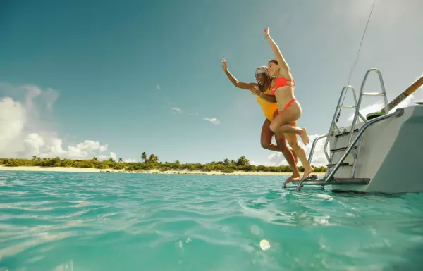 Two women in bright swimsuits jump joyfully from the back of a boat into clear turquoise ocean water near a tropical island shoreline in Puerto Rico under a sunny blue sky.