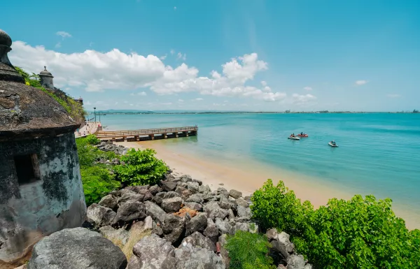 Panoramic coastal view from a historic fort in Old San Juan, Puerto Rico, overlooking turquoise water, a small beach, rocky shoreline, a wooden pier, and jet skis under a bright blue sky.